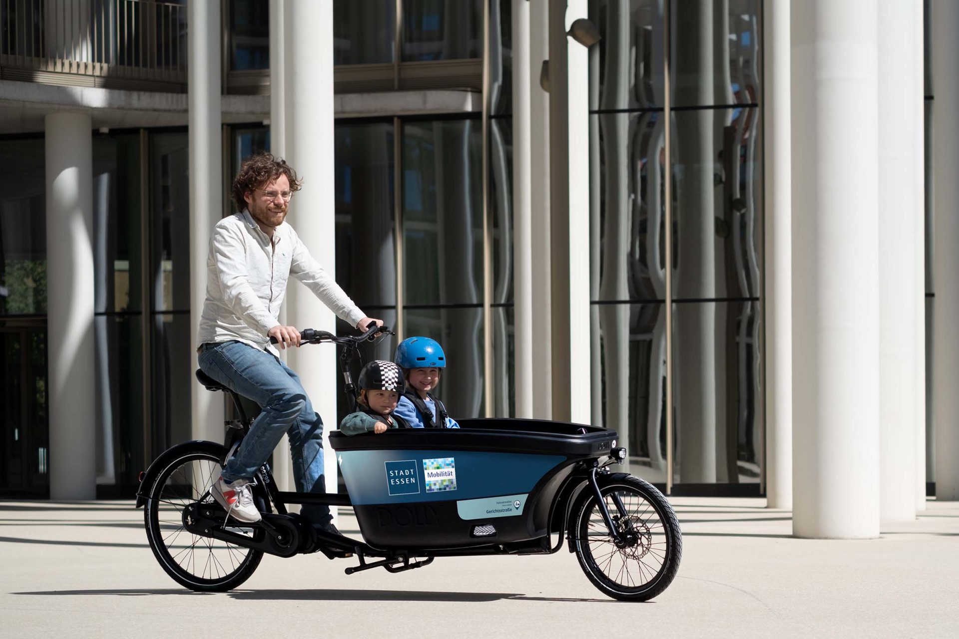 Ein Mann fährt ein StadtLastenRad Essen in dem zwei Kinder mit Helm sitzen.
